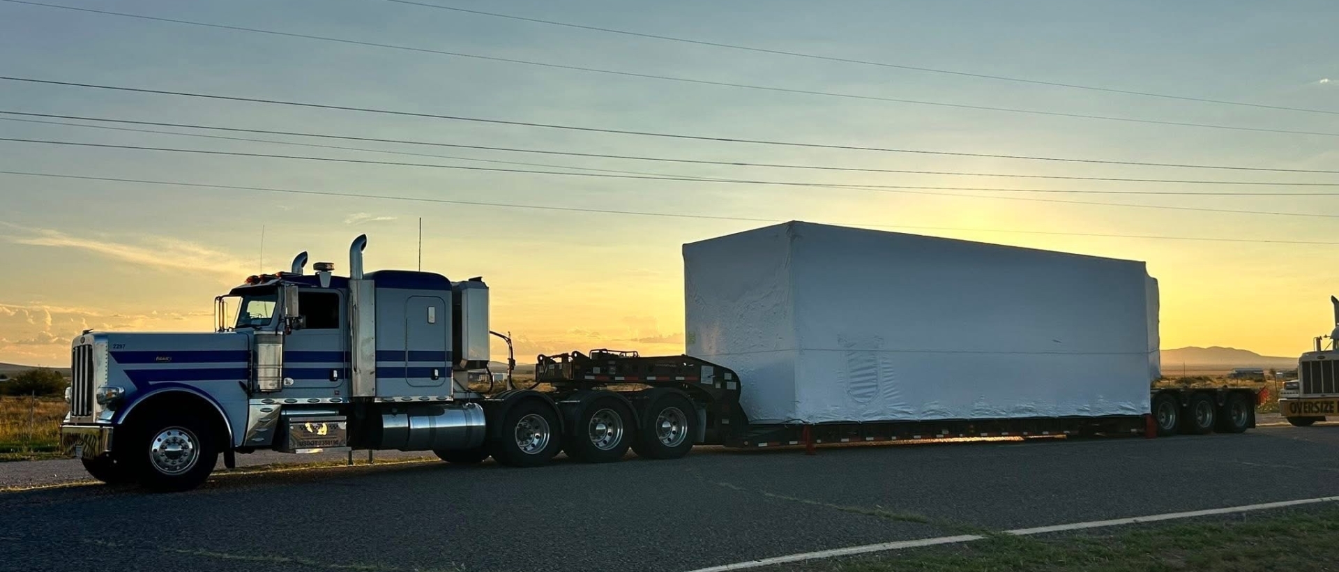 Powerful blue big rig semi truck with extended cab for truck driver rest transporting heavy haul cargo on flat bed semi trailer parked with the sunset in the background